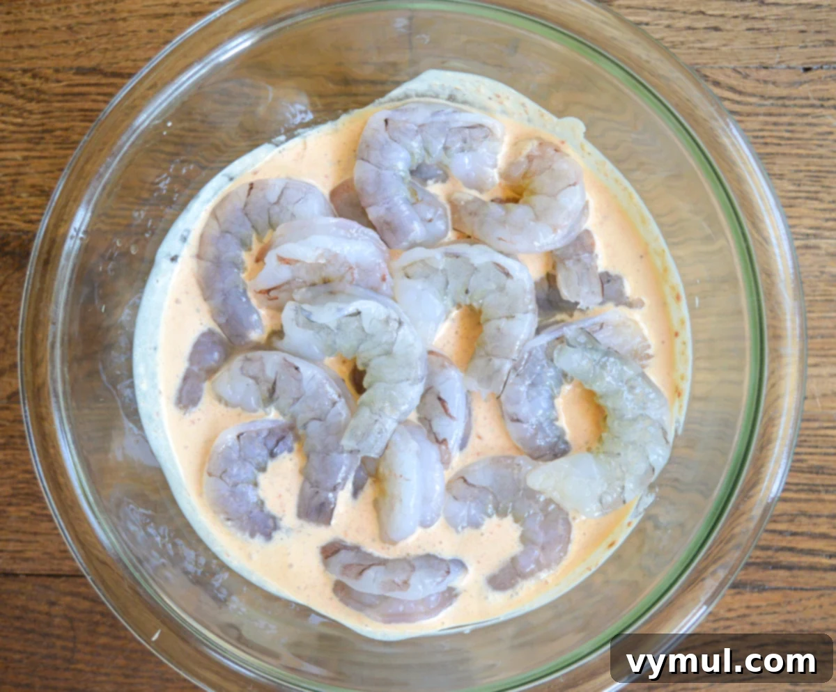 Raw shrimp marinating in a vibrant chipotle sauce within a clear glass bowl, preparing for grilling.
