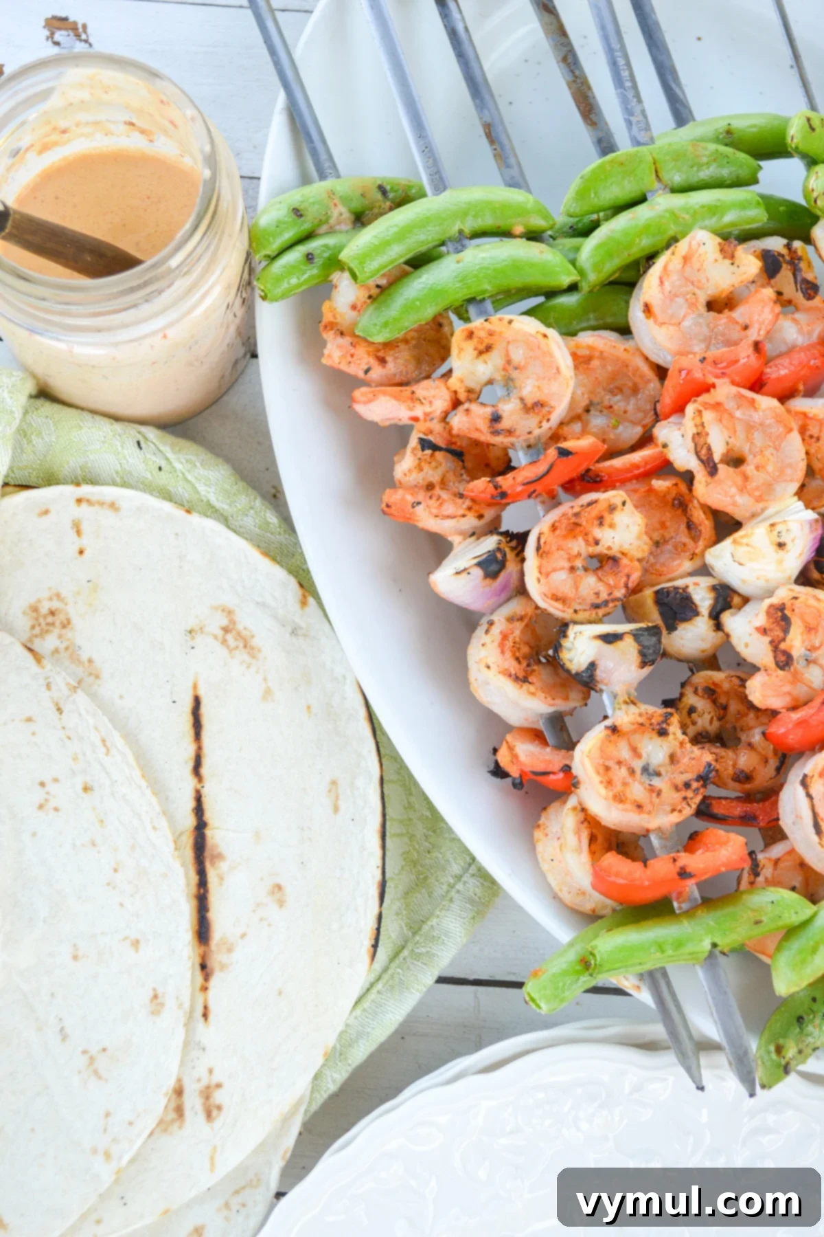 A beautiful spread of warm tortillas, a jar of creamy sauce, and a platter piled high with fresh grilled shrimp and vegetable skewers, ready for taco assembly.