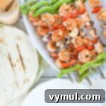 Close-up of a grilled shrimp and vegetable skewer, next to a small bowl of creamy chipotle sauce and warm tortillas.