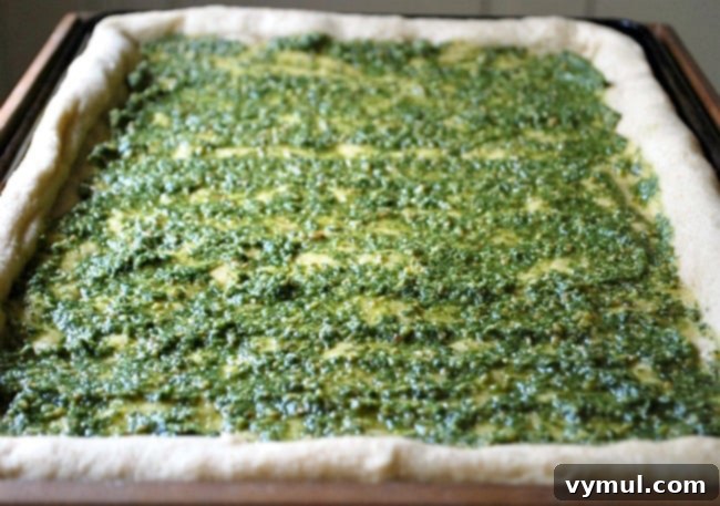 Spreading a vibrant green layer of homemade pesto sauce onto the pre-baked pizza dough for a Greek style pizza