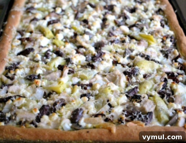 A freshly baked Greek style pizza with chicken, pesto, feta, and olives, hot out of the oven, golden brown and bubbly