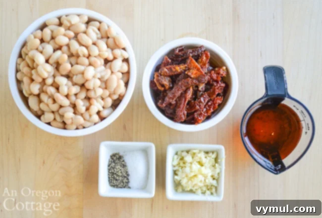 Ingredients for white bean dried tomato dip laid out on a table: white beans, sun-dried tomatoes, garlic, olive oil, salt, and pepper.