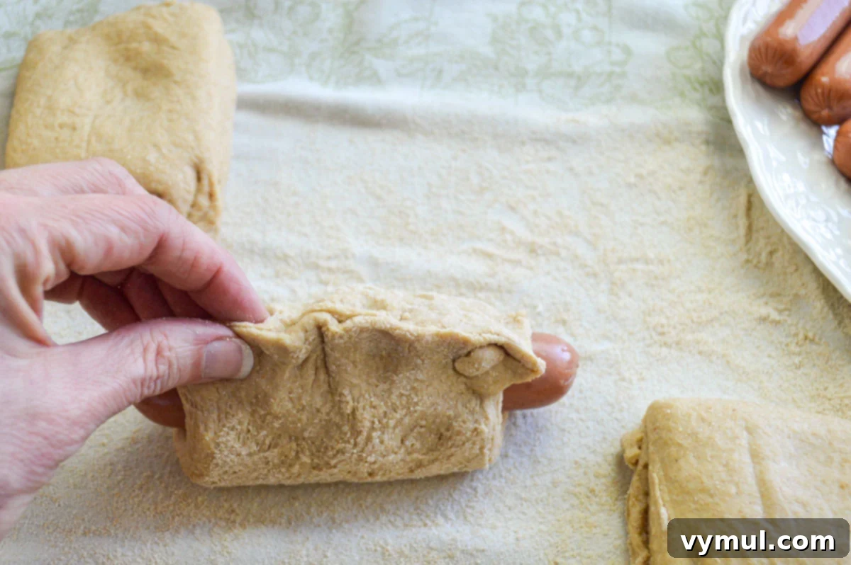 Easy 1-Hour Homemade Pigs In a Blanket 7 Hands pinching the edges of the homemade dough around a hot dog to seal it, forming a pigs in a blanket.