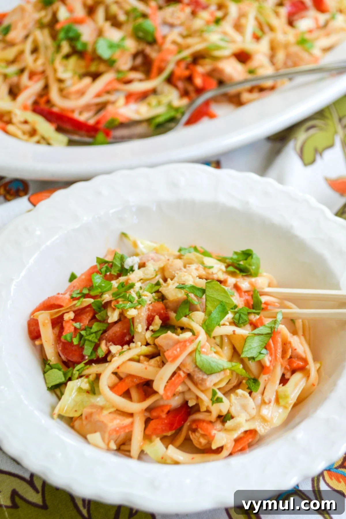 A pristine white bowl showcasing a generous serving of spicy peanut noodles, garnished with peanuts and fresh herbs, with chopsticks resting on the side.