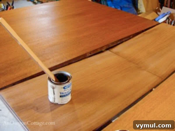 staining dining table-step6: A person applying wood stain to the dining table top with a foam brush, showing the color transformation.