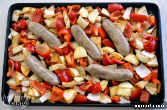 Sheet Pan Sausage & Veggies 5 Peppers and onions seasoned and spread on a baking sheet, with uncooked sausages nestled among them, ready for the oven