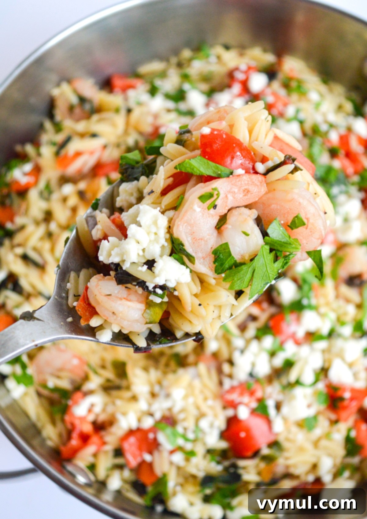 A serving spoon lifting a generous portion of shrimp orzo with vegetables and feta from a large skillet, ready to be enjoyed.