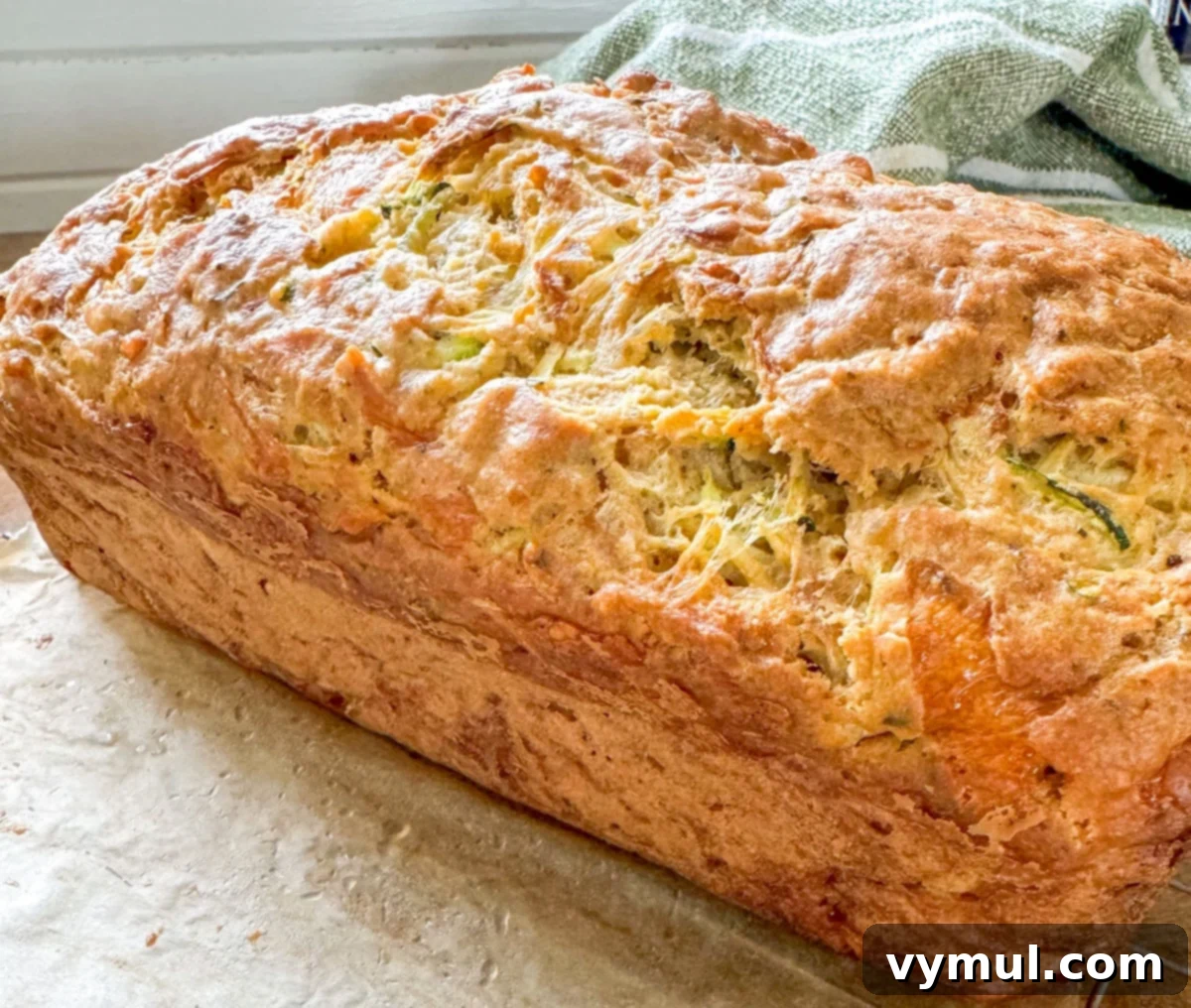 A golden-brown, baked loaf of savory zucchini bread, with a visibly crusty, cheesy top, resting out of the pan.