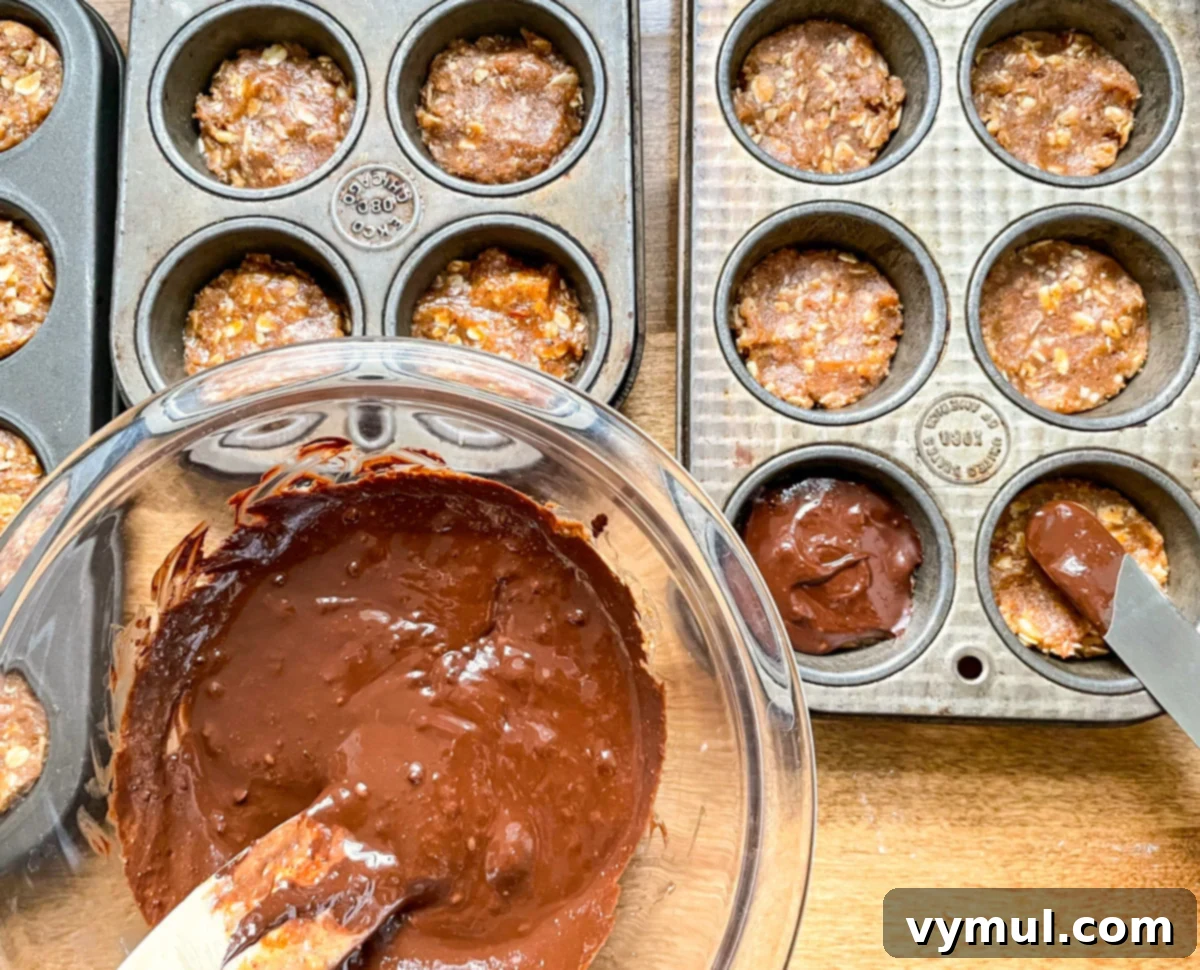Spreading smooth, melted chocolate topping evenly onto the frozen oat cup bases in a mini muffin tin using a spatula.