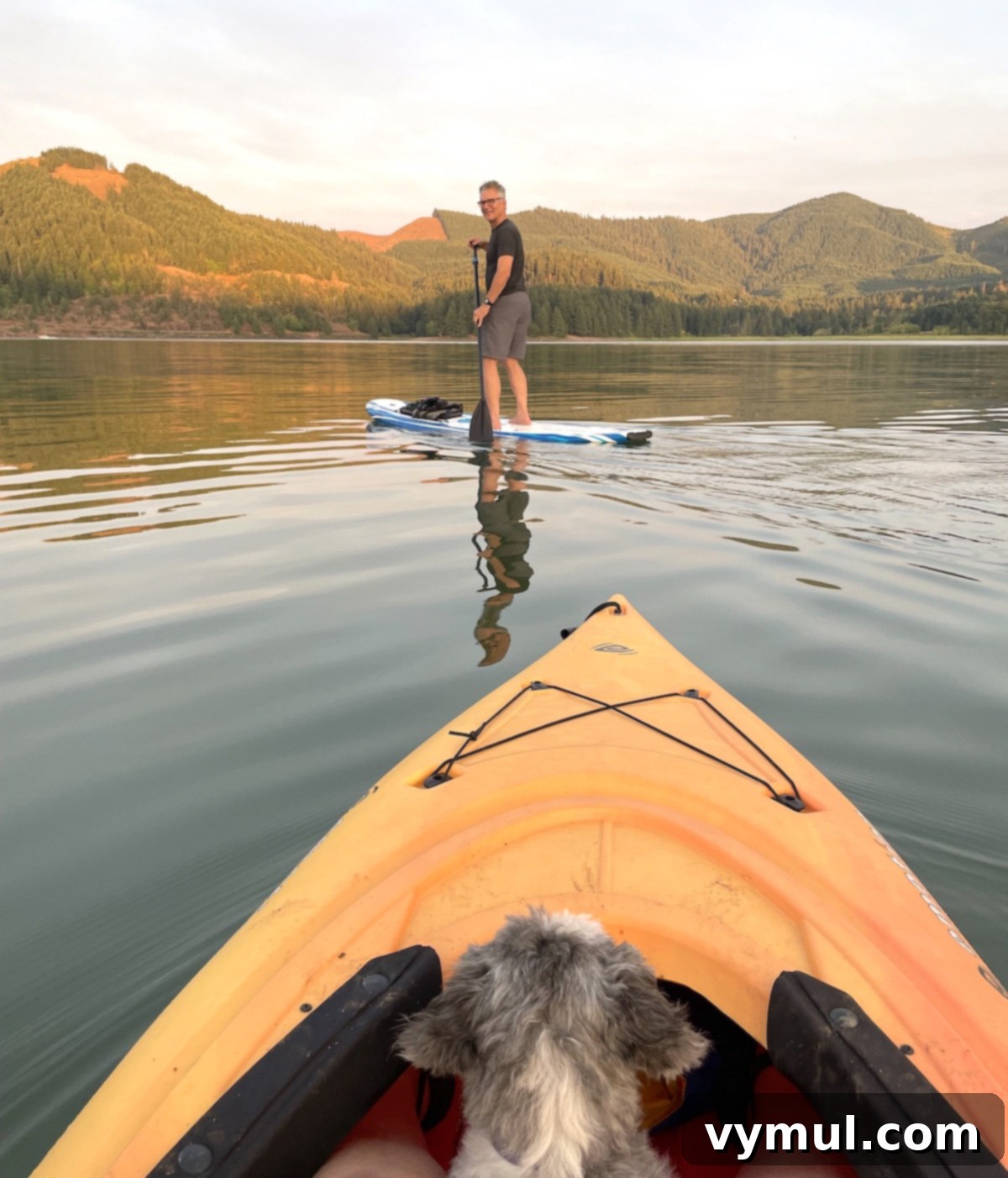 The 12 Gems of 2023 3 A serene scene of a person paddling a paddleboard on a calm lake, with another person in a kayak nearby. The sky is clear, reflecting on the water, and lush hills line the background. The focus is on peaceful outdoor activity.