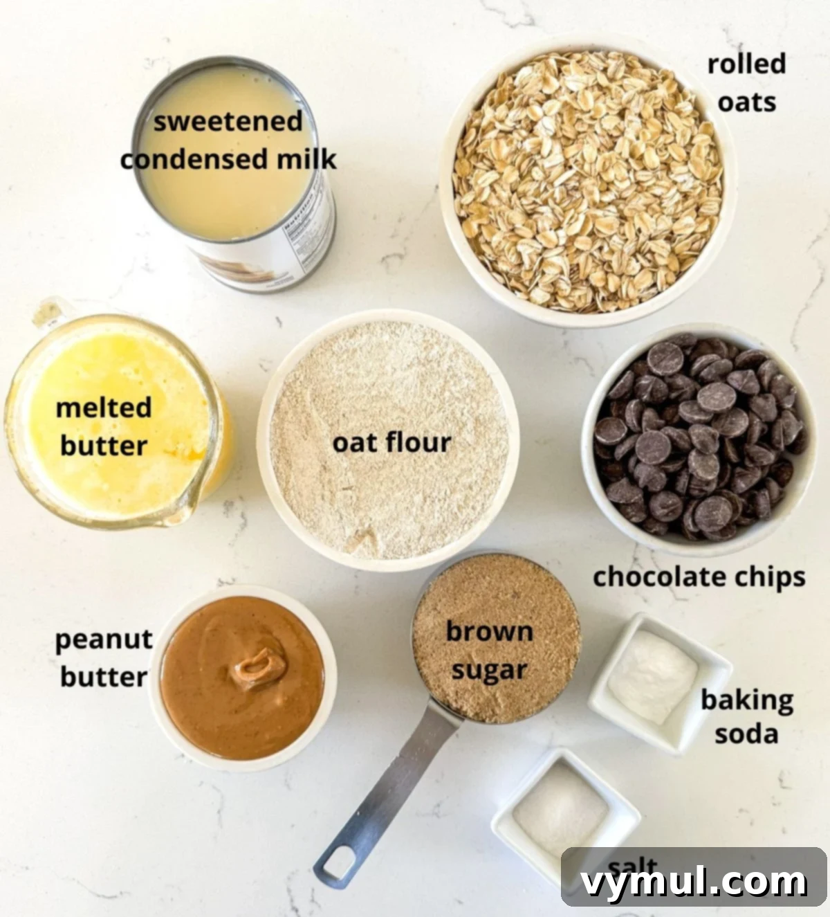 A collection of ingredients for peanut butter oat bars displayed on a marble counter, including oats, flour, peanut butter, condensed milk, and chocolate chips.