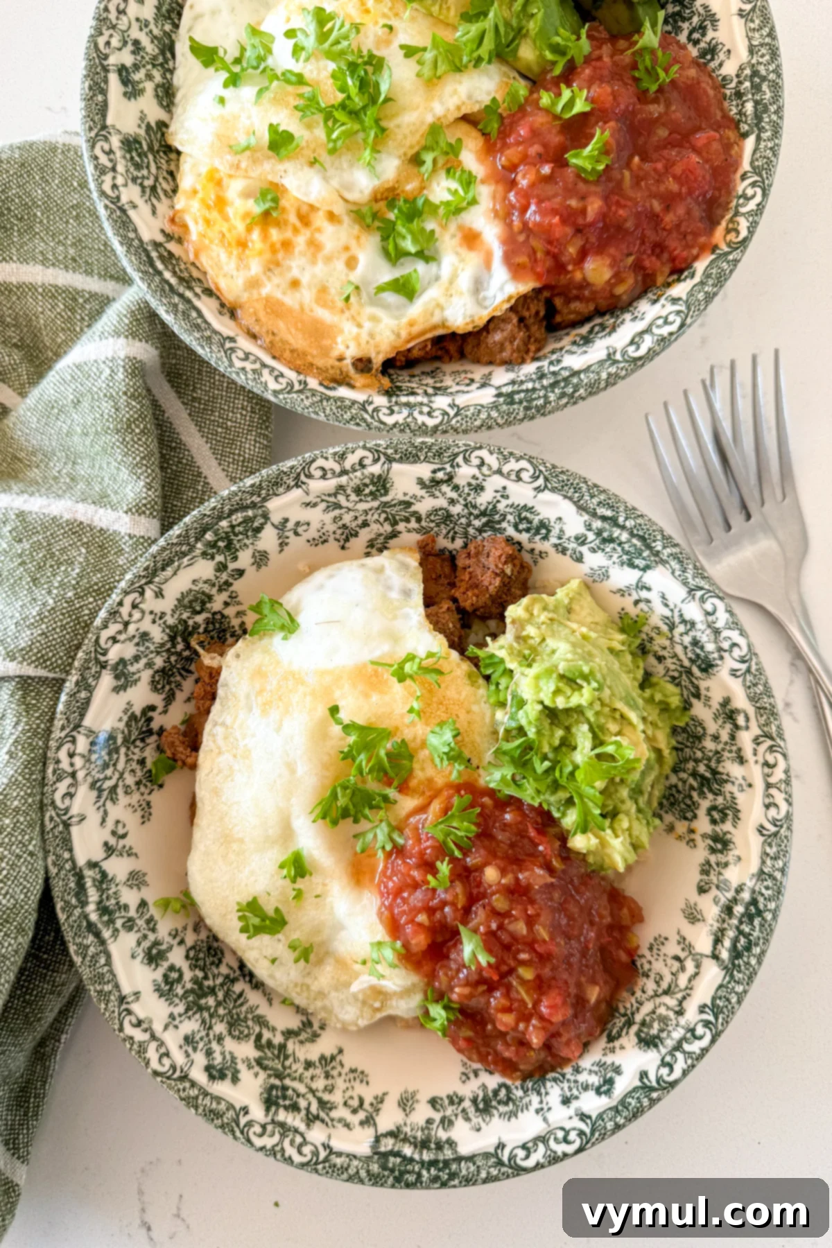 Speedy Six Ingredient High Protein Mexican Breakfast Bowls 2 Two vibrant Mexican breakfast bowls served in green floral dishes, viewed from above, showcasing eggs, ground meat, rice, avocado, and salsa.