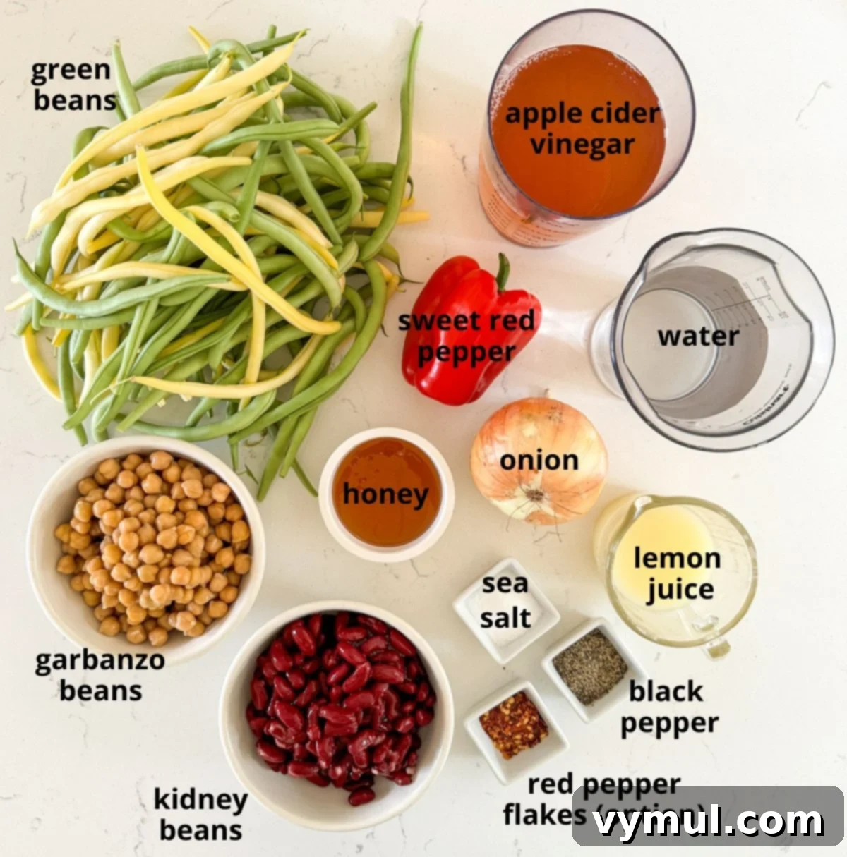 Assorted ingredients for three bean salad, including green beans, kidney beans, garbanzo beans, peppers, and onions, neatly arranged on a marbled counter.