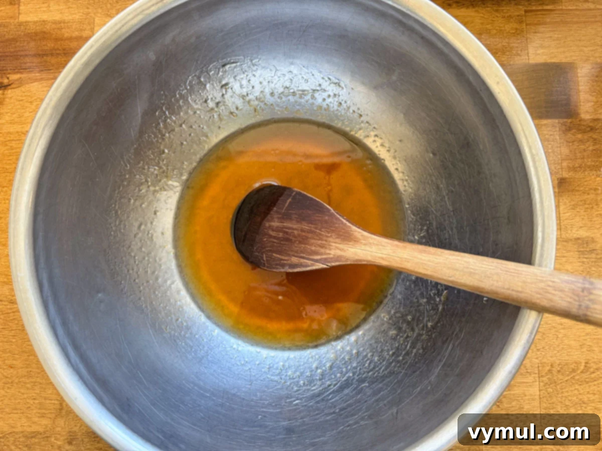 Wet ingredients for granola mixed in a large stainless steel bowl, showing the oil, syrup, and vanilla.