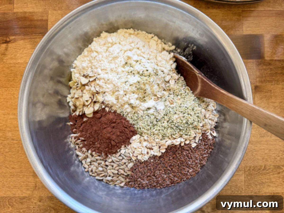 Dry granola ingredients, including oats, nuts, seeds, and protein powder, added to the wet mixture in a large stainless steel bowl and being stirred.