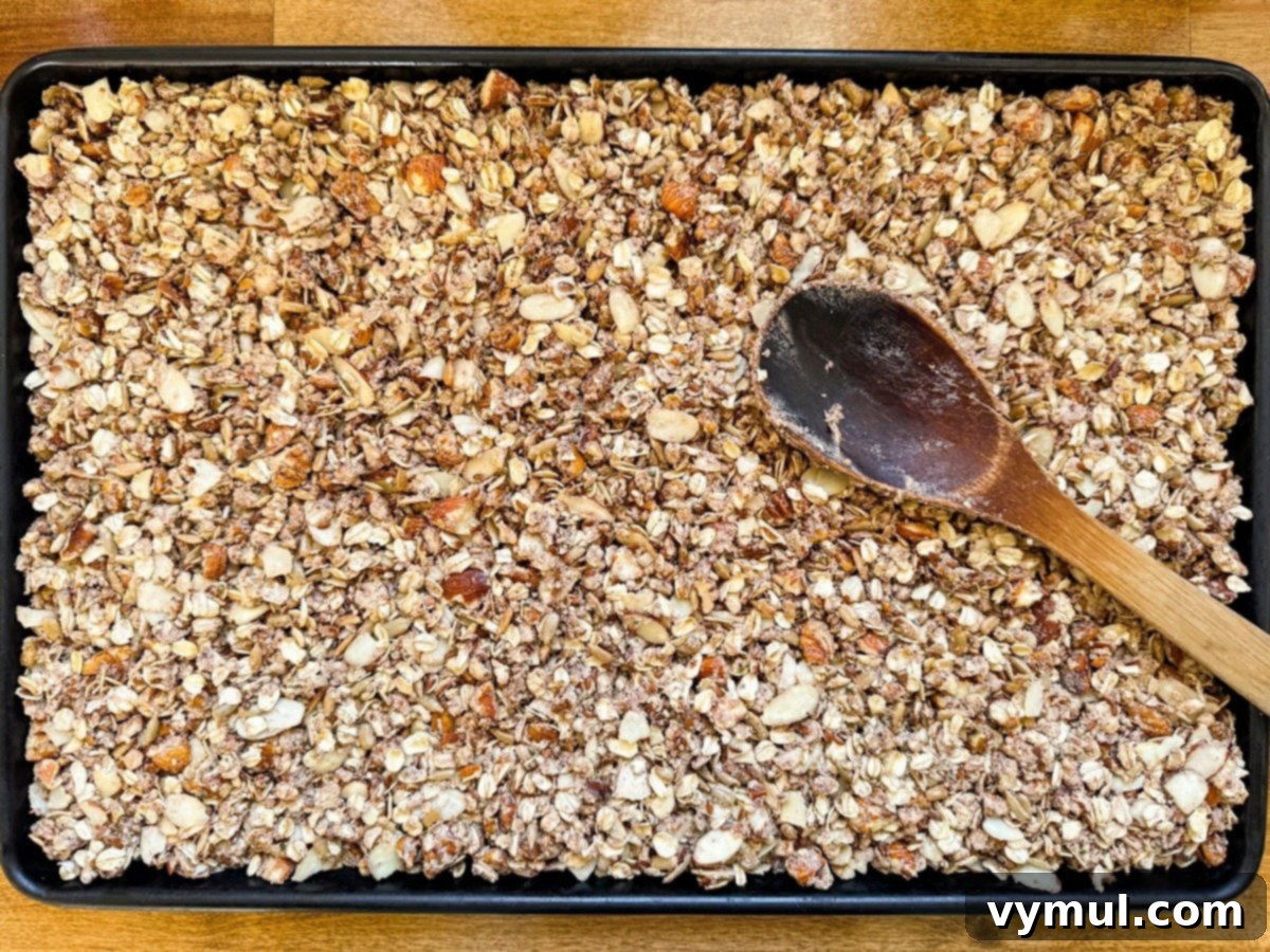 Granola mixture being patted flat and evenly onto a parchment-lined baking sheet using the back of a spoon.