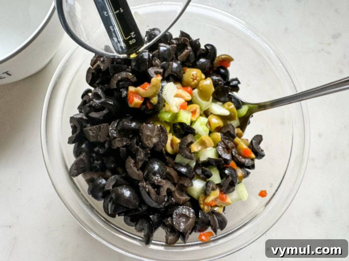 Effortless Olive Salad, Maximum Flavor 4 A glass bowl containing a vibrant mix of chopped olives, celery, and other ingredients, being tossed with olive oil and lemon juice, illustrating the simple preparation process for the olive salad.