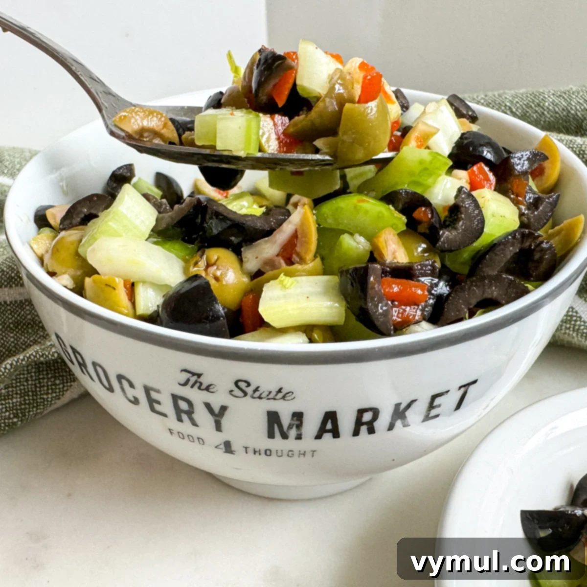 Effortless Olive Salad, Maximum Flavor 5 A white bowl filled with finished olive salad, garnished with fresh herbs, with a spoon poised to serve, highlighting the salad's readiness to be enjoyed.